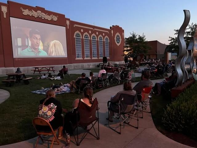 Outdoor movie night: people seated on chairs and blankets watching a film projected on a red brick wall, with an abstract sculpture nearby at dusk.