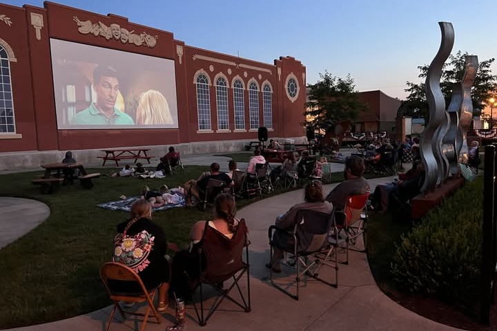 Outdoor movie night: people seated on chairs and blankets watching a film projected on a red brick wall, with an abstract sculpture nearby at dusk.