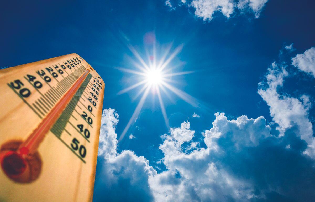 Wooden thermometer held up against a bright sun and blue sky with clouds, showing a high temperature on the scale (around 40–50).