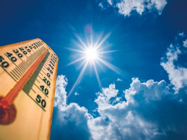 Wooden thermometer held up against a bright sun and blue sky with clouds, showing a high temperature on the scale (around 40–50).