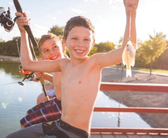 Two smiling boys on a dock with fishing rods, one proudly holding up a small fish.
