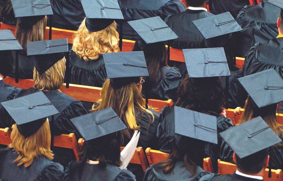 Rows of graduates in blue gowns and mortarboard caps seated in an auditorium with orange seats visible.
