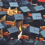 Rows of graduates in blue gowns and mortarboard caps seated in an auditorium with orange seats visible.