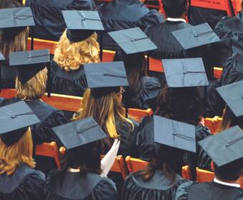 Rows of graduates in blue gowns and mortarboard caps seated in an auditorium with orange seats visible.