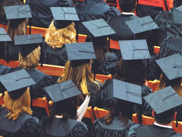 Rows of graduates in blue gowns and mortarboard caps seated in an auditorium with orange seats visible.