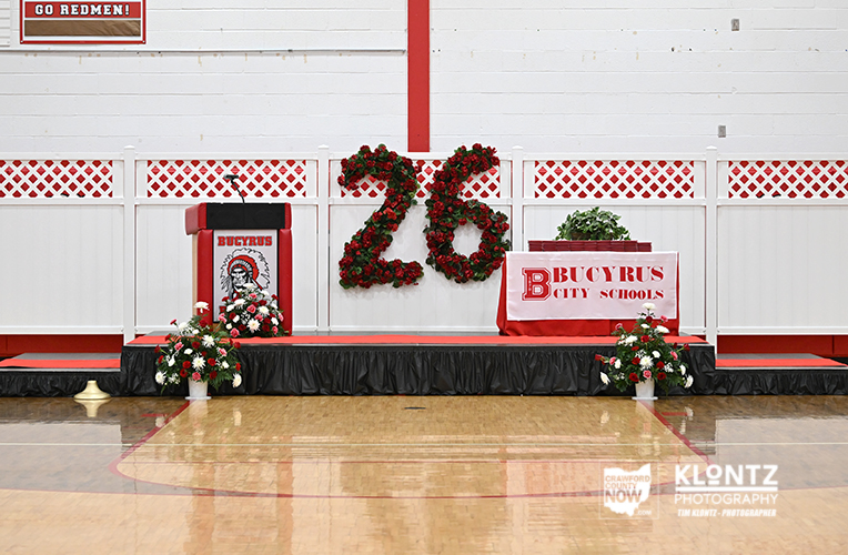 School gym stage decorated for a 26th anniversary with red-and-white banners, floral arrangements, and a Bucyrus City Schools sign on a table.