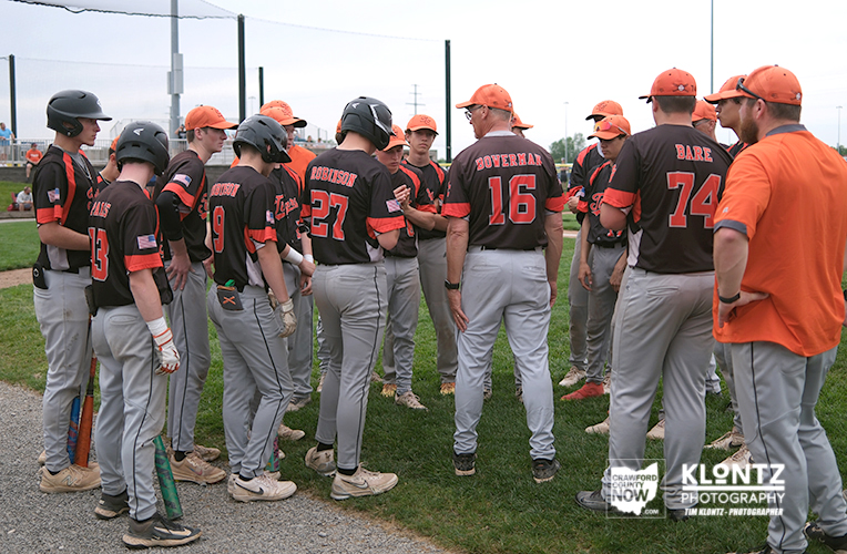 Baseball team in black and orange uniforms huddles on a grassy field, players wearing helmets while an adult coach speaks to them.