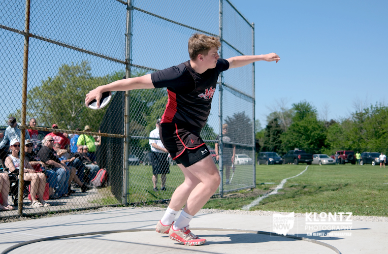 Young athlete in a black-red uniform throws a discus from a throwing circle near a chain-link fence with spectators watching.