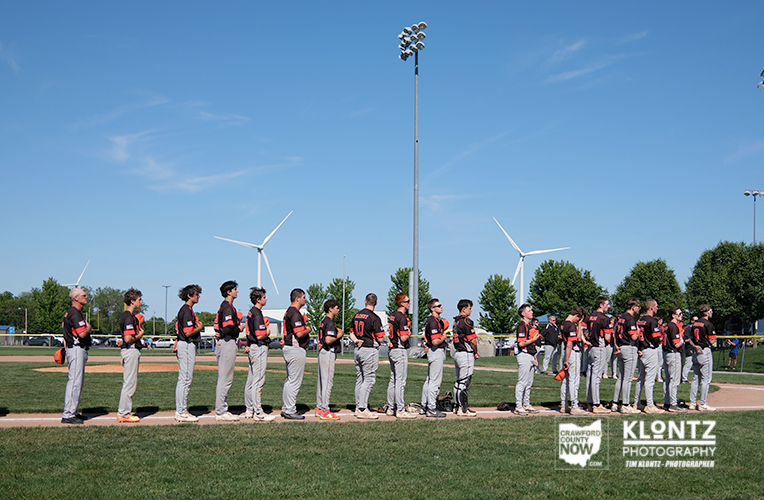 Line of baseball players in black and red uniforms stand in a row on the infield, facing left, with wind turbines and a tall stadium light pole against a clear blue sky behind them.