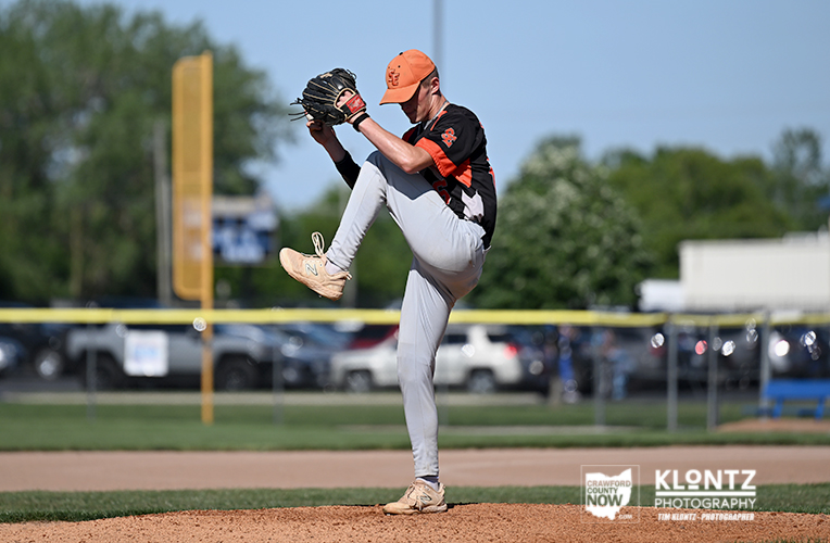 Baseball pitcher in an orange cap and black jersey delivering a wind-up on the mound, raised leg mid-throw.