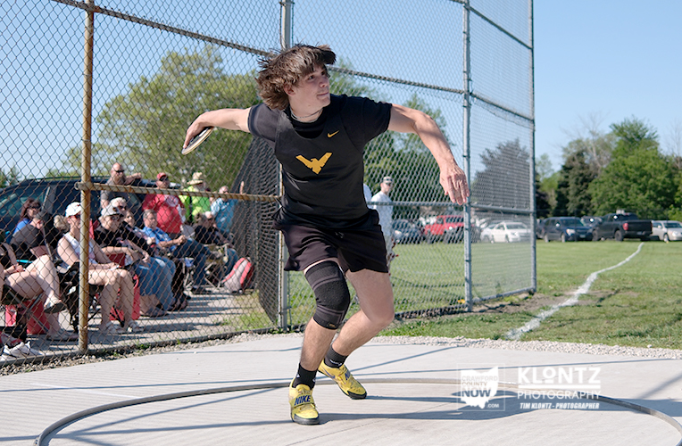 Male athlete in a black shirt and knee brace mid-throw with a discus inside a throwing circle, spectators seated behind a chain-link fence on a sunny day.