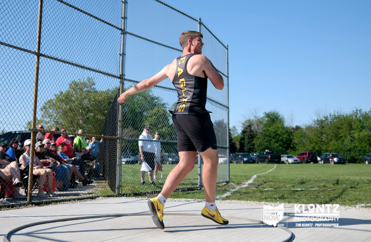 Male athlete in a black and yellow singlet pivots in a throwing circle during a discus event, with spectators behind a chain-link fence on a sunny day.