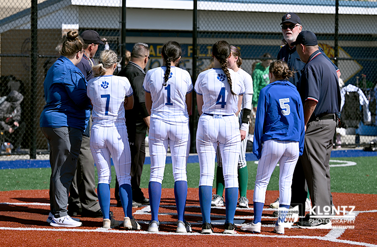 Girls softball team huddled in a circle on the infield, backs to camera, white uniforms with blue numbers.