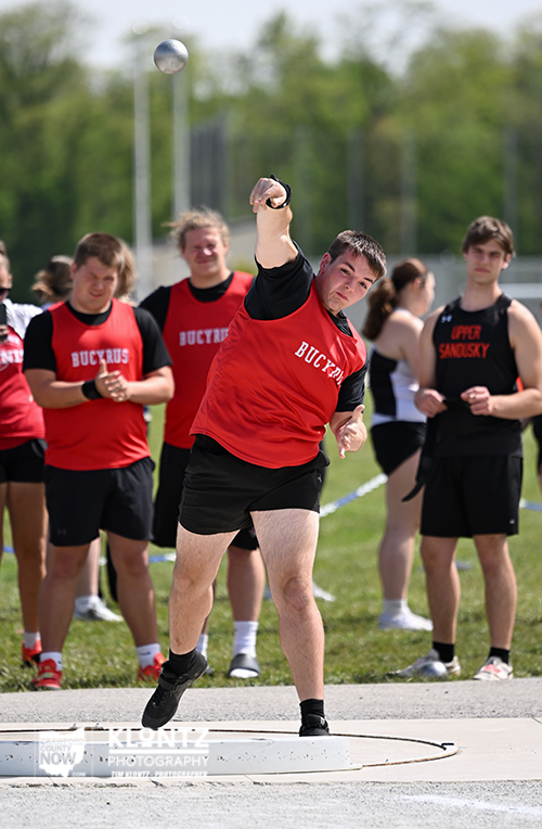Male shot-putter in a red singlet and black shorts mid-throw, with teammates watching on a field in the background.