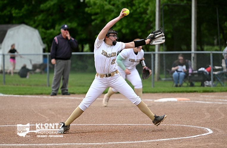 Softball pitcher in a white uniform delivering a pitch on the dirt field, ball in her raised hand.