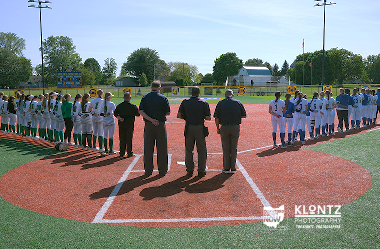 Two youth softball teams line up along the baselines on a sunny field before a game, with four officials standing at home plate.