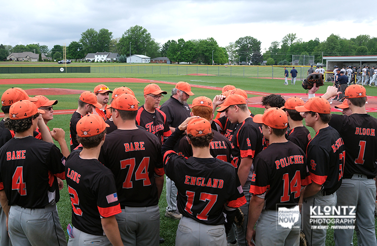 A group of baseball players in black uniforms with orange caps huddle on a baseball field during practice or a game.