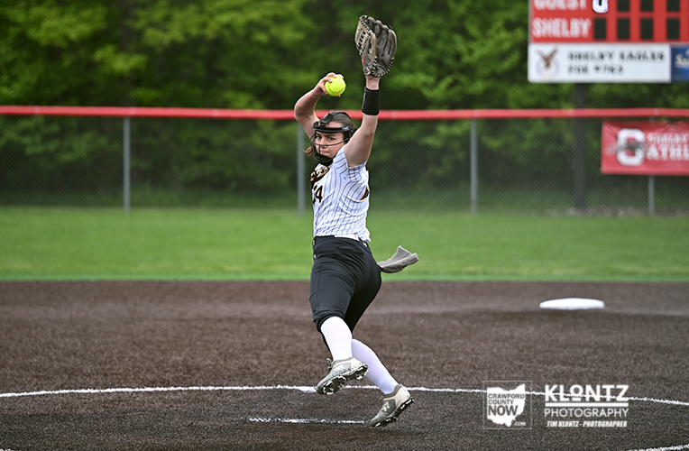 Female softball pitcher in mid-pitch on the mound, throwing a yellow ball with glove raised, wearing a striped jersey and black pants.