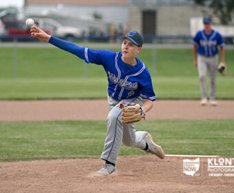 Youth baseball pitcher in a blue uniform throws a pitch from the mound, with a fielder in the background on the grass field.