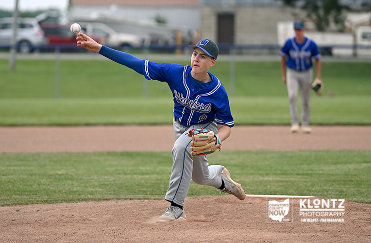 Youth baseball pitcher in a blue uniform throws a pitch from the mound, with a fielder in the background on the grass field.