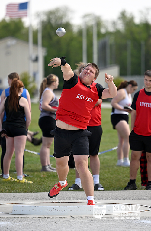 Male athlete in a red singlet throws a shot put during a track and field field event, with spectators watching on a grassy area in the background.