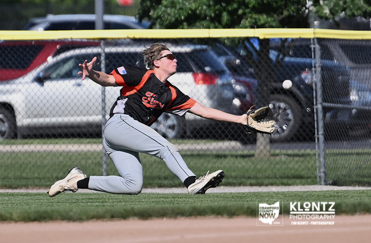 Baseball fielder diving to the left, reaching with glove to catch a ball on the dirt infield under a chain-link fence.