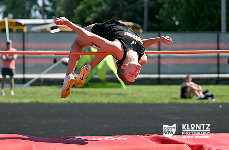 Female high jumper in black clears an orange bar over a red padded mat at a track stadium.