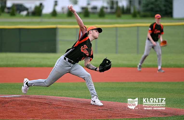 Baseball pitcher in orange and black uniform delivering a pitch from the mound as a fielder watches from the infield in the background