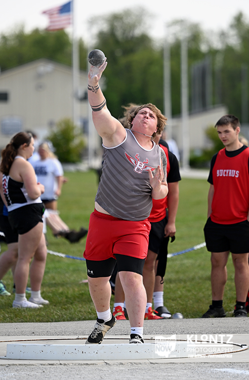 Athlete in gray shirt and red shorts throws a shot put on a track field, with teammates and a flag in the background.