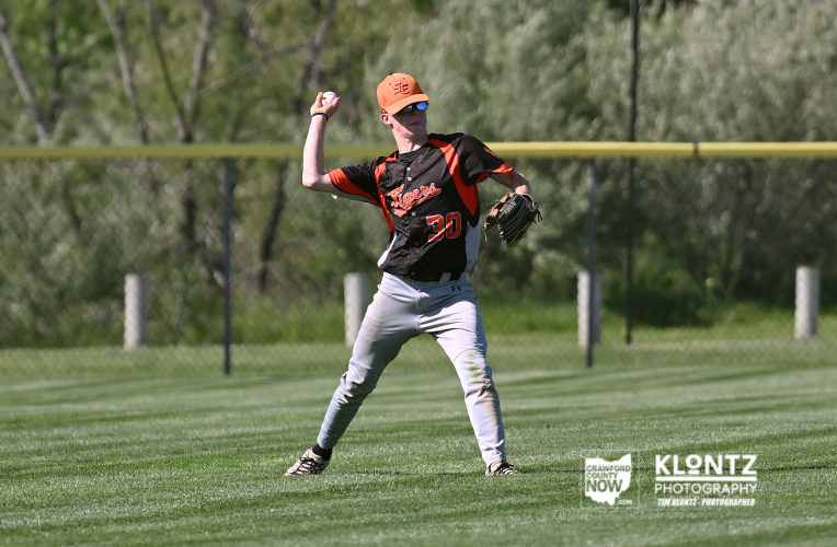 Baseball pitcher in black and orange uniform throwing a ball on a grassy field with a yellow outfield fence behind him.