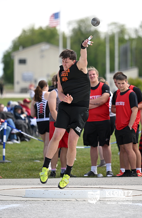 Male track athlete in a black uniform mid-throw of the shot put during a competition, with teammates and a flag in the background.
