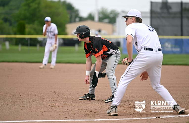 Young fielder in black and orange crouches at a base, ready to field as a baserunner advances.