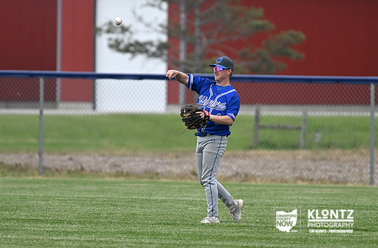 Baseball fielder in a blue uniform throws a ball mid-air, glove ready, on a grassy field with a chain-link fence in the background.