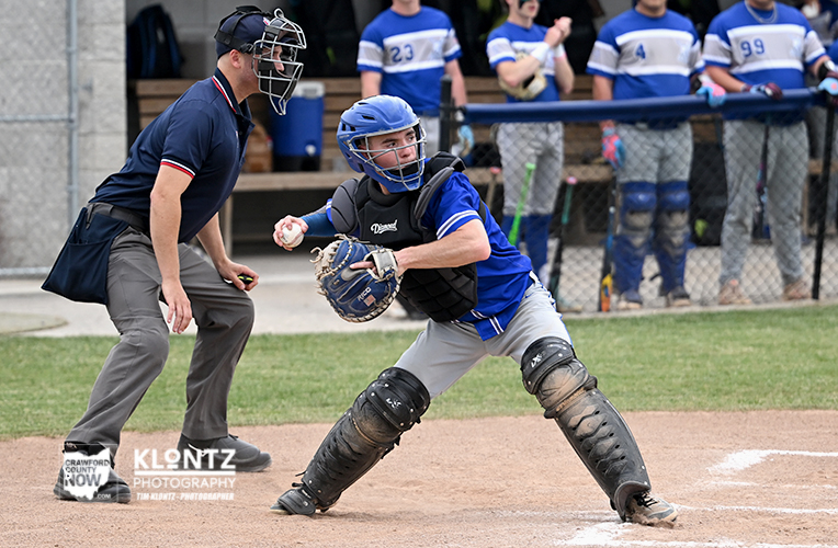 Baseball catcher in blue with helmet and protective gear at home plate, ready to tag, as an umpire watches and teammates in blue stand in the background.