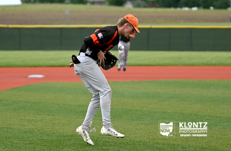 Baseball player in an orange cap and black jersey crouches to field a ball on a green baseball diamond.