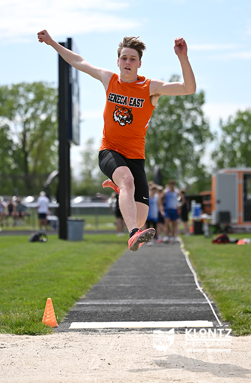 Male athlete in an orange Genese East jersey mid-air while performing a long jump at a track meet, sand pit ahead and cones along the runway.