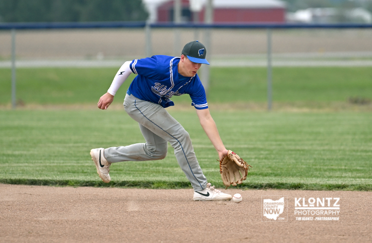 Baseball player in a blue jersey fields a ground ball, stretching to reach it on the dirt infield.