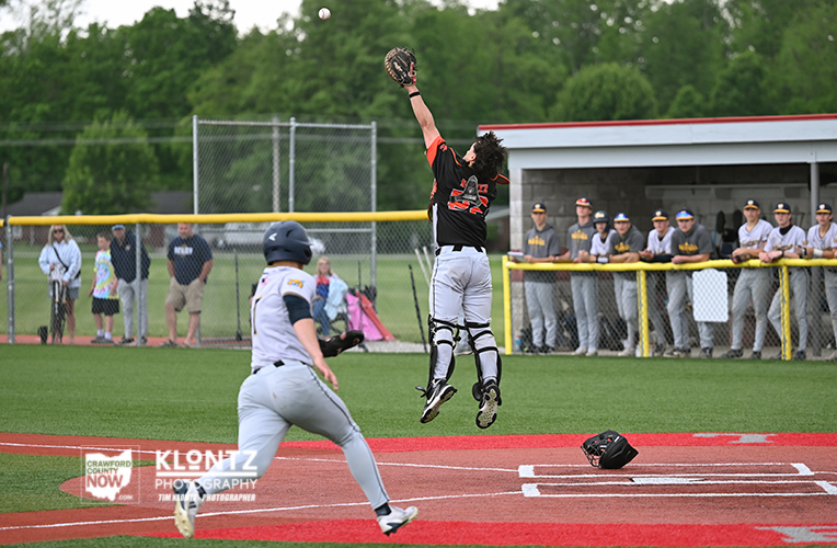 Baseball catcher in black jumps to catch a high throw at home plate, pitcher running toward the base in the foreground with spectators in the background.