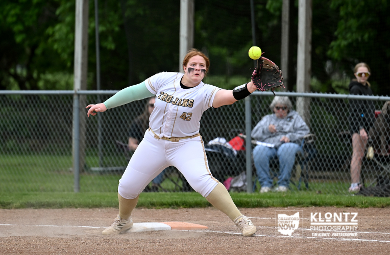 Female softball pitcher in a white Trojans uniform (#42) delivering a pitch on the infield dirt, glove outstretched as a bright ball approaches the glove.