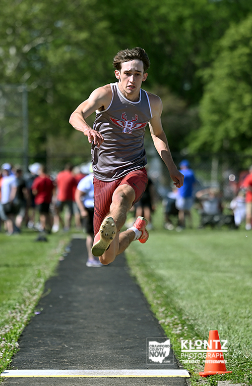 Male athlete in a gray sleeveless shirt and red shorts leaps mid-air in a long jump on a track, with spectators and greenery in the background.