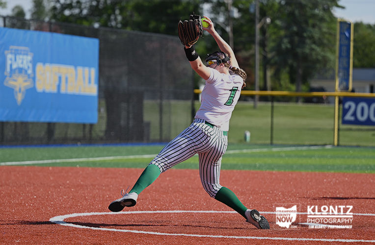 Softball pitcher in a white uniform with green socks mid-pitch on the mound.