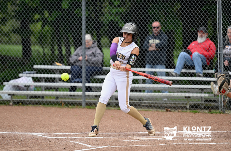 Female softball batter in a white uniform swings at a pitched ball at home plate, wearing a helmet and purple sleeve.