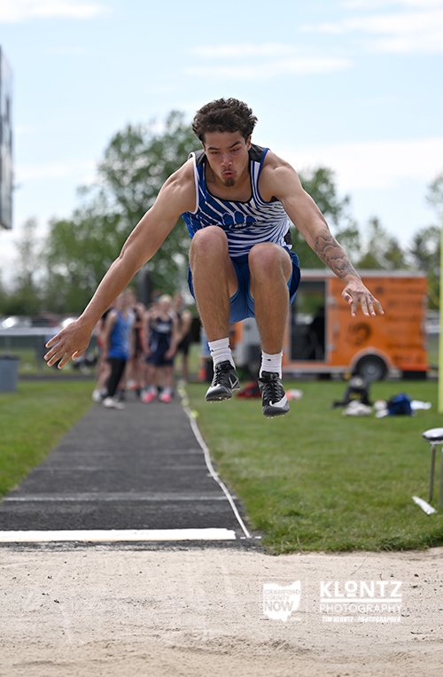 Male athlete in a blue sprint uniform leaps mid-air during a long jump at an outdoor track, with the sand pit and spectators in the background.