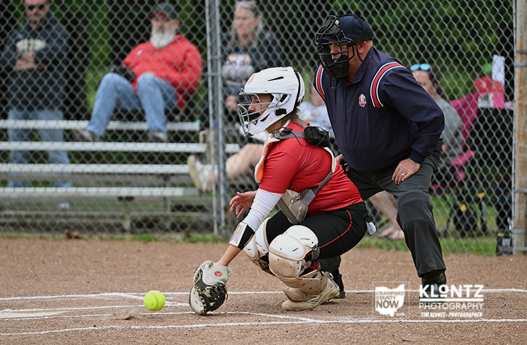 Catcher in red gear crouches at home plate in protective gear as a green softball rolls toward her glove, umpire behind her.