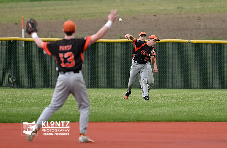 Baseball players in orange and black uniforms on a grassy field; one fielder with number 13 faces away in the foreground while two teammates watch a throw in midair near the outfield fence.