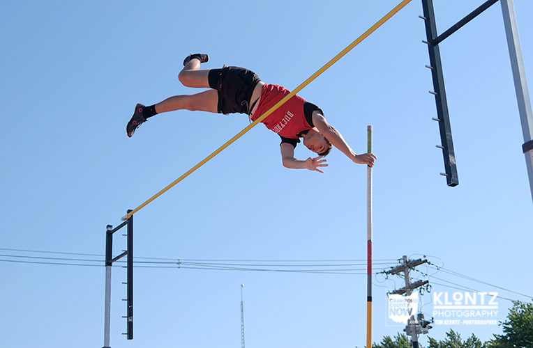 Male athlete in a red sleeveless shirt and black shorts clears a high jump bar, upside down mid‑flight against a clear blue sky.