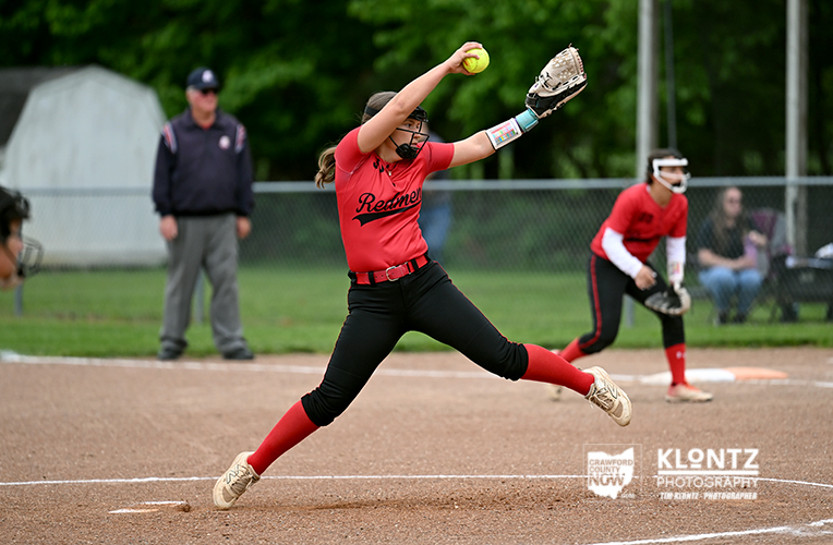 Softball pitcher in red uniform mid-pitch on the mound, ball raised in her throwing hand as a catcher and umpire watch nearby.