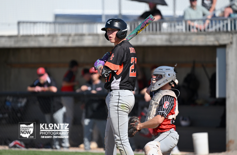 Baseball batter in black-and-orange uniform stands at home plate with a colorful bat, wearing a helmet, as the catcher in red and white gear guards behind him.