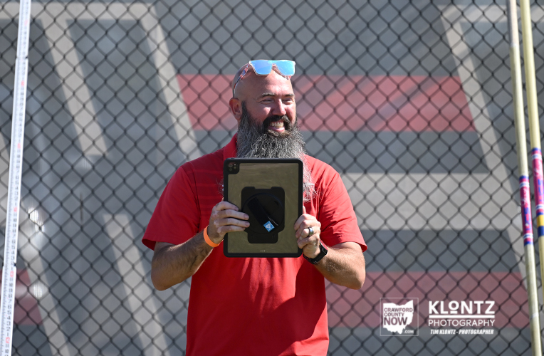 Bearded man in a red shirt holding a tablet, smiling outdoors in front of a chain-link fence.