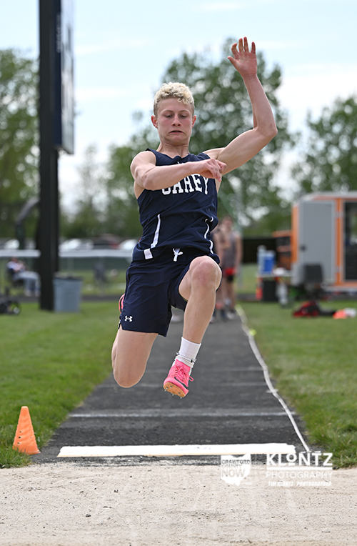 Female long jumper in a navy uniform mid-air over the takeoff board during a track and field event with a sand pit ahead.